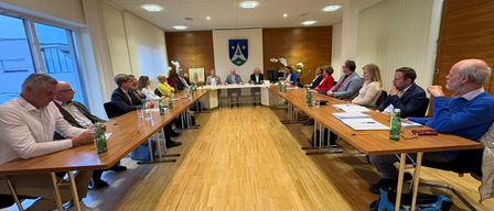 Eine Besprechung im Gange mit Teilnehmern, die um einen Konferenztisch in einem Raum mit Holzboden und einer mit einem Wappen geschmückten Wand sitzen.