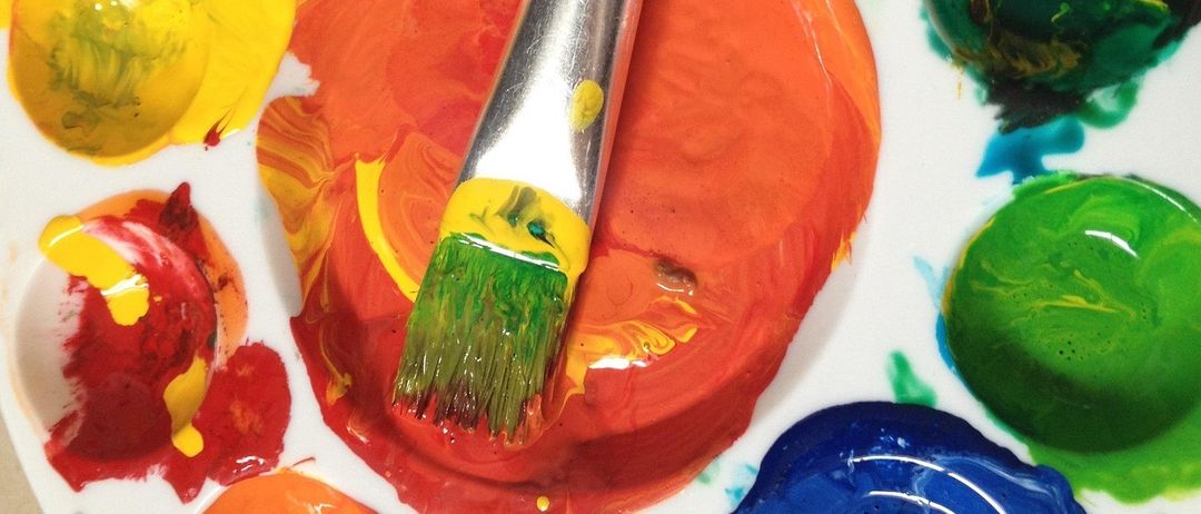 Eine weiße Palette mit verschiedenen Farben in unterschiedlichen Bereichen und einem in orangefarbene Farbe getauchten Pinsel.