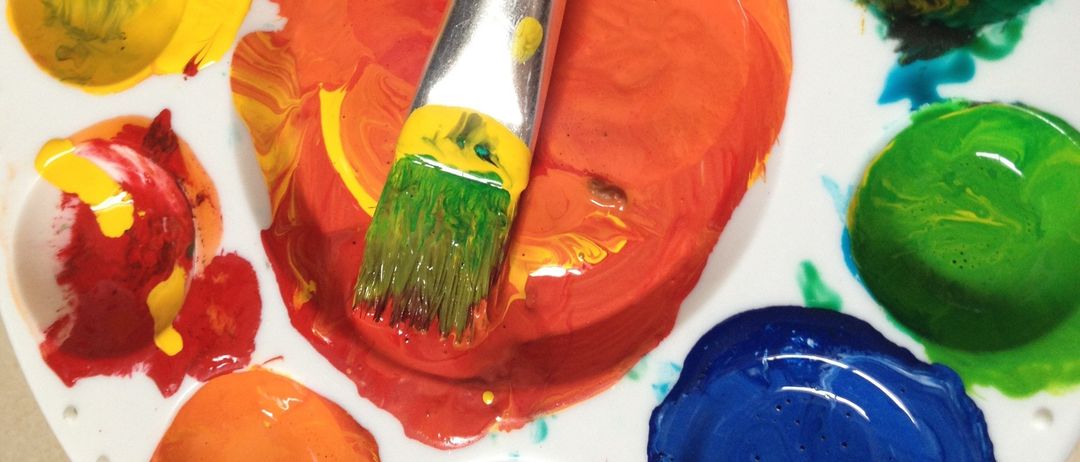 Ein weißes Malbrett mit verschiedenen farbigen Farben in den Mulden, einschließlich Rot, Blau, Gelb und Grün. Ein Pinsel mit mehrfarbiger Farbe liegt auf dem Malbrett.