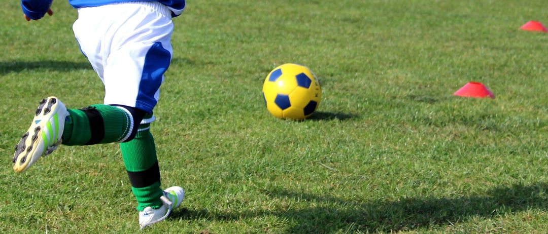 Ein junger Junge, der ein blaues und weißes Fußballtrikot trägt, läuft auf dem Feld. Der Ball liegt auf dem Feld und ein Torpfosten ist hinter ihm.