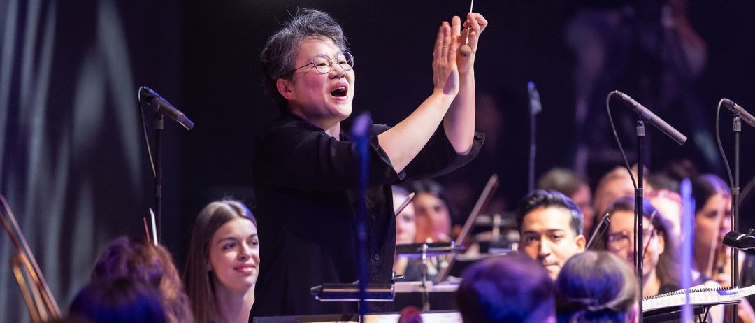 Ein Dirigent steht vor einem Orchester, hält einen Taktstock und singt, während das Orchester spielt. Eine Frau in der vorderen Reihe schaut lächelnd zu.