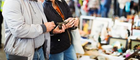 Ein Mann und eine Frau betrachten ein kleines, kompliziertes Objekt auf einem Flohmarkt, umgeben von verschiedenen ausgestellten Gegenständen.