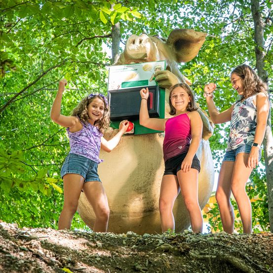 Drei junge Mädchen stehen neben einer großen Schweineskulptur im Wald und posieren für ein Foto. Das Schwein hält eine Schachtel und die Mädchen lächeln.