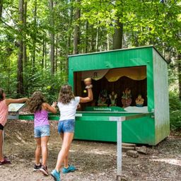 Drei Mädchen spielen ein Spiel mit Bällen im Wald, in der Nähe einer grünen Kabine mit einem Schild, das 'Fehlerbehebung' steht.