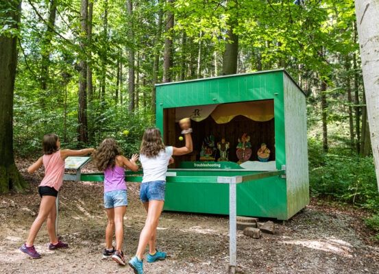 Drei Mädchen spielen ein Spiel mit Bällen im Wald, in der Nähe einer grünen Kabine mit einem Schild, das 'Fehlerbehebung' steht.