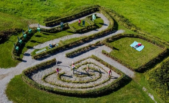 Eine Draufsicht eines Parks mit einem gewundenen Pfad und einem Spirallabyrinth. Mehrere Menschen gehen auf dem Pfad, während andere auf dem Gras sitzen oder liegen.