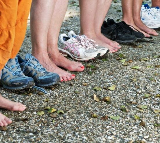 Fünf Personen stehen barfuß auf einem Kiesweg, jeder mit seinen Schuhen in der Nähe. Die Schuhe sind in verschiedenen Farben und Stilen.
