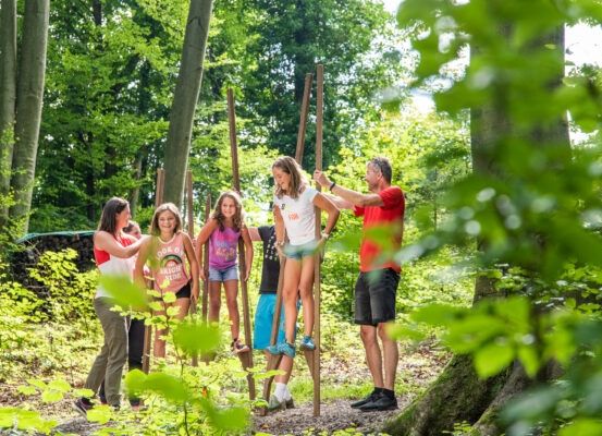Eine Gruppe von Kindern und Erwachsenen spielt im Wald. Sie stehen auf Holzpfählen, einige tragen Turnschuhe.