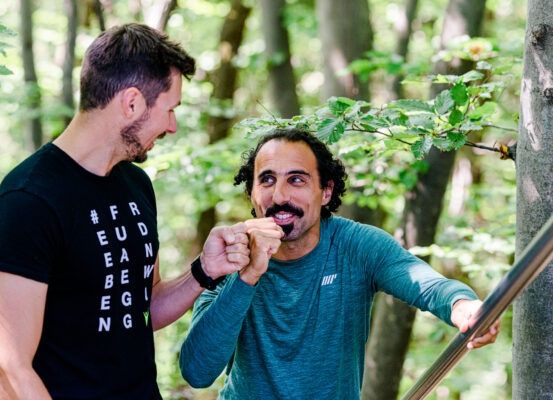 Zwei Männer im Wald; einer lächelt, hält einen Baum fest, während der andere eine Faust macht.