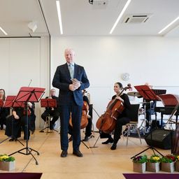Ein Mann in einem Anzug steht vor einem Orchester und hält eine Musikpartitur. Die Musiker sitzen und spielen verschiedene Instrumente. Die Szene spielt in einer Halle mit einer schlichten weißen Wand im Hintergrund.