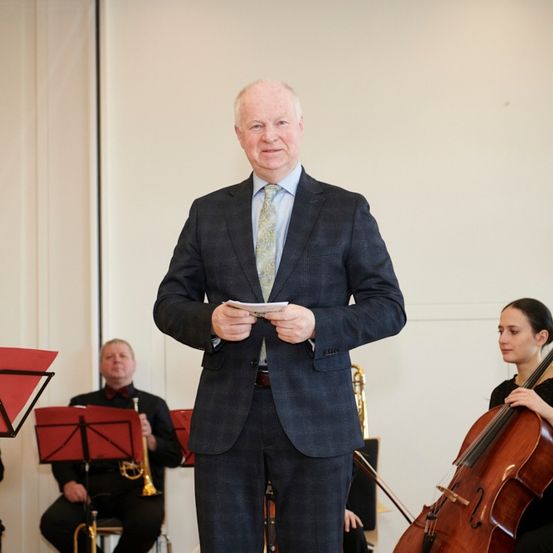 Ein Mann in einem Anzug hält ein Stück Papier in der Hand, während er vor einer Gruppe von Musikern steht, die sitzen und verschiedene Instrumente spielen.