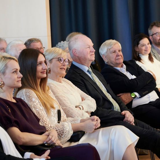 Eine Gruppe von Menschen in formeller Kleidung sitzt bei einer Veranstaltung in Stühlen. Sie scheinen in Gespräche vertieft zu sein. Hinter ihnen ist ein blauer Vorhang zu sehen.