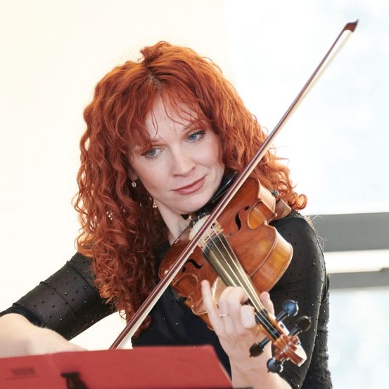 Eine Frau mit lockigem rotem Haar spielt Geige. Sie trägt ein schwarzes Oberteil und lächelt, während sie auf die Geige schaut. Sie hält die Geige mit der linken Hand und den Bogen mit der rechten Hand.