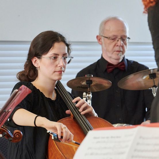 Eine Frau spielt Cello, während ein älterer Mann mit Brille und Fliege zusieht und hinter einem Schlagzeug steht.