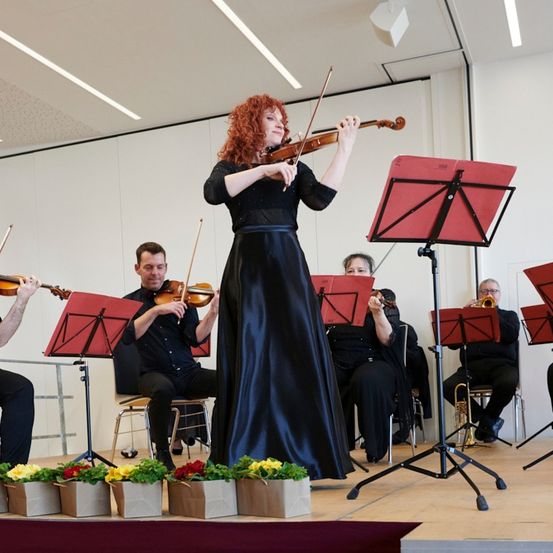 Eine Frau in einem schwarzen Kleid spielt Geige auf einer Bühne mit einem kleinen Orchester, umgeben von roten Notenständern und Blumen.
