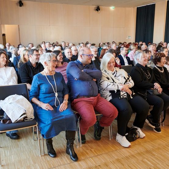 Ein großes Publikum sitzt in Reihen in einem modernen Raum, mit Brillen und Freizeitkleidung. Eine Frau in einem blauen Kleid und eine andere in einer weißen und schwarzen Jacke sitzen in der vorderen Reihe.