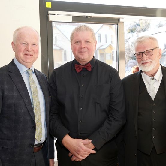Drei Männer stehen dicht beieinander in der Nähe einer Glastür. Der Mann auf der linken Seite trägt einen karierten Anzug, der Mann in der Mitte ein schwarzes Hemd und eine Fliege und der Mann auf der rechten Seite einen schwarzen Anzug und eine Brille. Hinter ihnen steht ein Gebäude mit gelber Fassade.