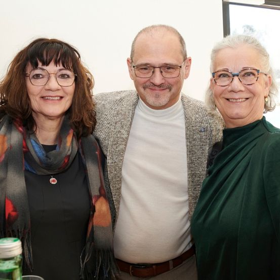 Drei Menschen, zwei Frauen und ein Mann, lächeln und posieren für ein Foto. Die Frau auf der linken Seite trägt eine Krawatte und eine Brille. Der Mann in der Mitte trägt eine Brille und einen Gürtel. Die Frau auf der rechten Seite trägt Ohrringe und ein grünes Oberteil.