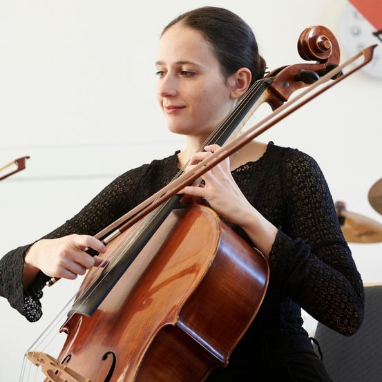 Eine Frau in einem schwarzen Oberteil spielt Cello und konzentriert sich auf ihre Aufführung in einem Musikraum.