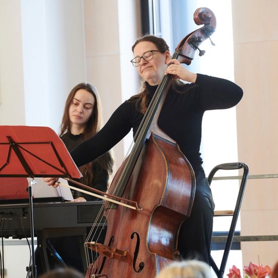 Eine Frau in Schwarz spielt Cello, während eine andere Frau Klavier spielt. Sie treten in einem Raum mit einem roten Notenständer und Publikum auf.