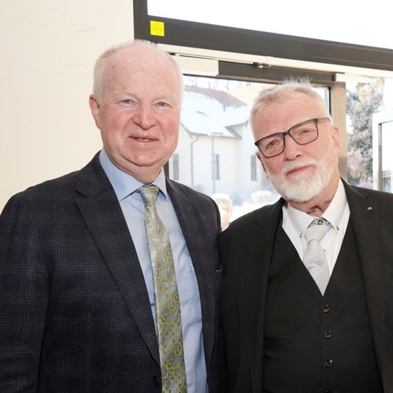 Zwei Männer in Anzügen stehen nebeneinander und lächeln. Einer trägt eine Krawatte mit Paisleymuster, der andere hat einen weißen Bart und Brille. Hinter ihnen ist ein Glasfenster.