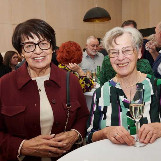 Zwei Frauen, beide mit Brille, sitzen bei einer Veranstaltung an einem Tisch. Die Frau auf der linken Seite trägt eine weinrote Jacke. Die Frau auf der rechten Seite hält ein Weinglas.
