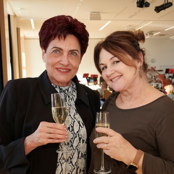 Zwei Frauen lächeln und halten Champagnerflöten. Die Frau links hat kurzes rotes Haar und trägt einen schwarzen Mantel. Die Frau rechts hat einen Dutt und trägt ein braunes Hemd.