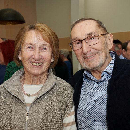Ein älteres Paar steht eng beieinander und lächelt für ein Foto, mit verschwommenen Menschen im Hintergrund.