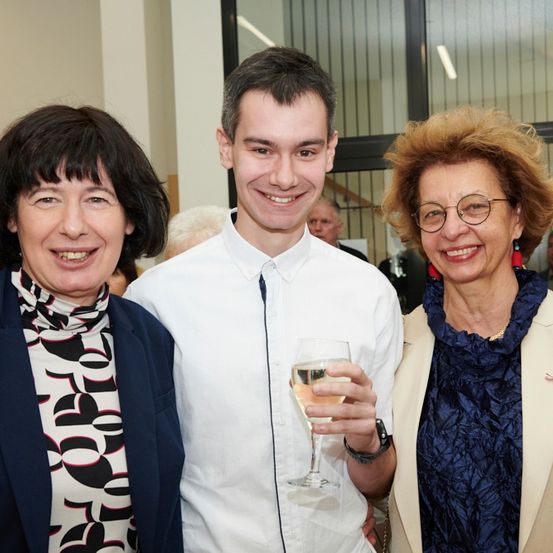 Drei Personen lächeln für ein Foto; ein Mann in der Mitte mit einem Weinglas und zwei Frauen auf beiden Seiten. Hinter ihnen sind verschwommene Figuren zu sehen.