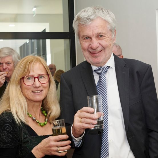 Ein Mann und eine Frau lächeln und halten Gläser mit Getränken, wahrscheinlich bei einer gesellschaftlichen Veranstaltung. Hinter ihnen stehen mehrere Personen, einige essen.