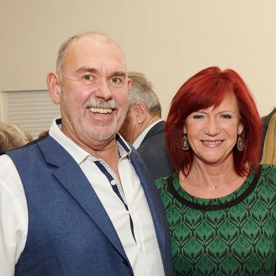 Ein älterer Mann und eine Frau lächeln und posieren für ein Foto. Der Mann trägt ein blaues Weste und weißes Hemd, und die Frau hat rote Haare und ein grün gemustertes Oberteil.