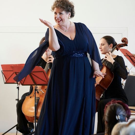 Eine Frau in einem blauen Kleid steht lächelnd auf der Bühne, mit einem Cellisten und einer weiteren Musikerin hinter ihr, vor einem Publikum.