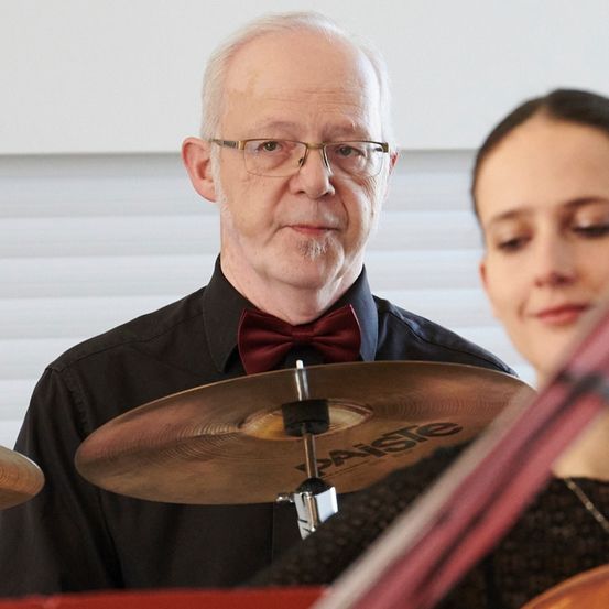 Ein älterer Mann mit Brille und roter Fliege spielt eine Becken, während eine Frau eine Geige spielt. Sie sind in einem Raum mit weißen Jalousien.