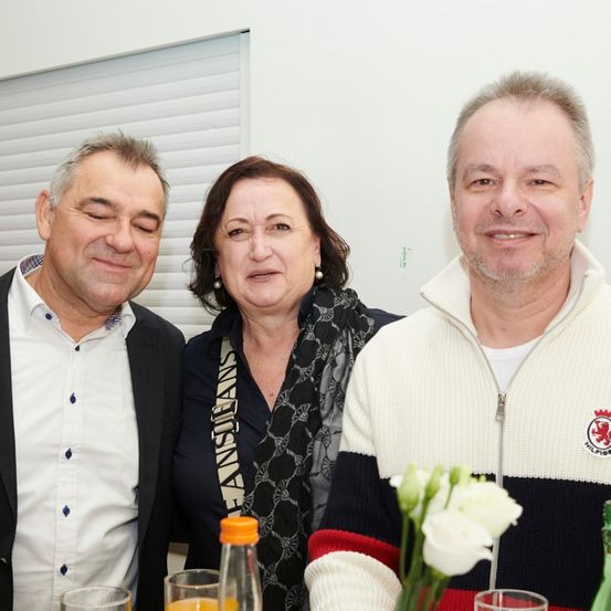 Drei Personen, zwei Männer und eine Frau, lächeln und posieren für ein Foto bei einer Veranstaltung. Sie stehen eng beieinander, mit einer weißen Blumenarrangement und Flaschen vor ihnen.