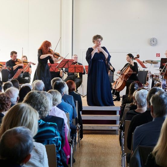 Eine Gruppe von Musikern tritt auf der Bühne vor einem sitzenden Publikum auf. Geiger, Cellisten und ein Trompeter sind anwesend. Die Frau im blauen Kleid scheint zu singen.