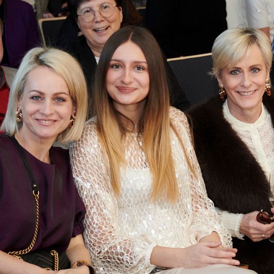 Drei Frauen sitzen zusammen und lächeln. Die Frau auf der linken Seite trägt ein lila Hemd. Die Frau in der Mitte hat lange blonde Haare. Die Frau auf der rechten Seite hält ein Glas Wein.