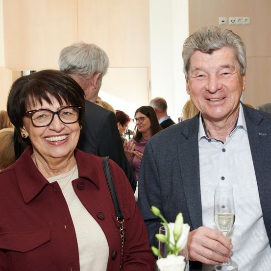 Ein lächelndes Paar bei einer Veranstaltung, die Frau mit Brille und einem burgunderroten Mantel, der Mann im Anzug mit einem Weinglas. Hinter ihnen unterhalten sich verschiedene Personen.