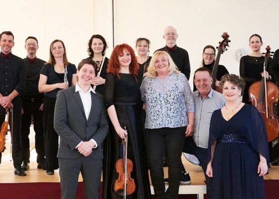 Eine Gruppe von Musikern posiert für ein Foto auf einer Bühne, einige halten Musikinstrumente in der Hand, lächeln und andere stehen hinter ihnen.