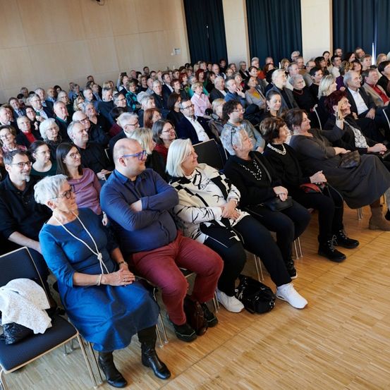 Eine große Menschenmenge sitzt in einem Raum, einige tragen Brillen und Halsketten. Der Raum hat Holzböden und blaue Vorhänge.