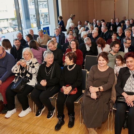 Eine große Menschenmenge, überwiegend ältere Erwachsene, sitzt in Stühlen in einem Raum. Einige benutzen ihre Handys, während andere nach vorne schauen. Hinter ihnen sind Autos vor dem Gebäude geparkt.