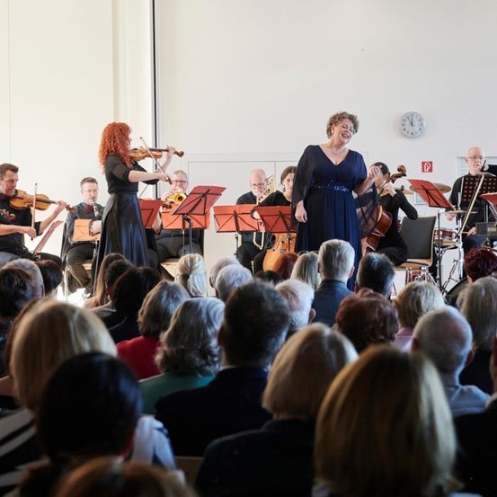 Eine Gruppe von Musikern spielt auf der Bühne mit Violinen, Celli, Trompeten und Klavier. Ein Publikum beobachtet von vorne, wobei eine Uhr und ein Ausgangsschild an der Wand zu sehen sind.