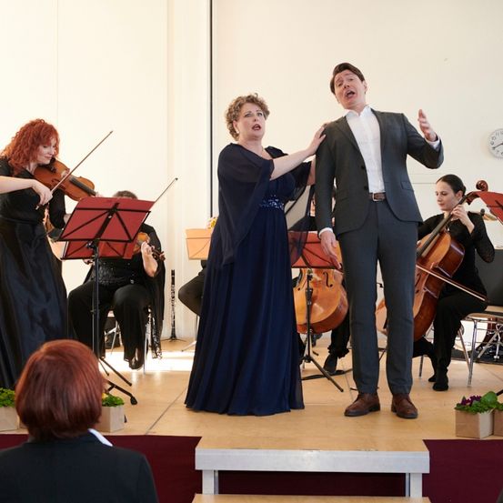 Ein Mann und eine Frau spielen mit einem Streichquartett auf der Bühne. Eine Geigerin steht hinter dem Mann. Hinter ihnen sind Cellisten. Vor der Bühne stehen Blumentöpfe.