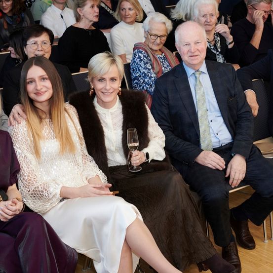 Eine Gruppe von Menschen, darunter ein Mann und zwei Frauen, sitzt zusammen in einem Innenraum. Die Frau in der Mitte hält ein Weinglas, und der Mann neben ihr trägt eine Krawatte. Sie alle scheinen zu lächeln.