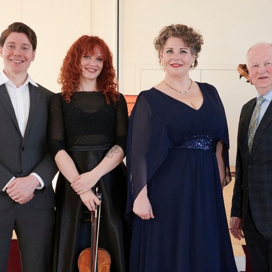 Vier Personen in formeller Kleidung stehen lächelnd zusammen. Ein Mann links hält eine Geige, während eine Frau neben ihm eine Geige und einen Cello hat. Eine weitere Frau steht neben ihr, und ein Mann ist rechts.