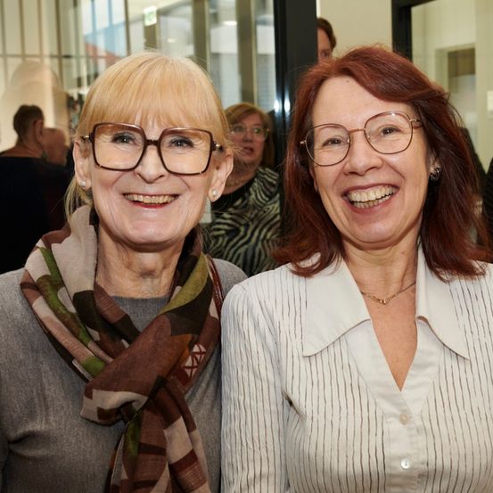 Zwei Frauen, beide mit Brille, stehen eng nebeneinander und lächeln. Eine trägt ein Halstuch, die andere eine Halskette. Sie scheinen sich bei einer Veranstaltung zu befinden, wobei die Menschen im Hintergrund verschwommen sind.
