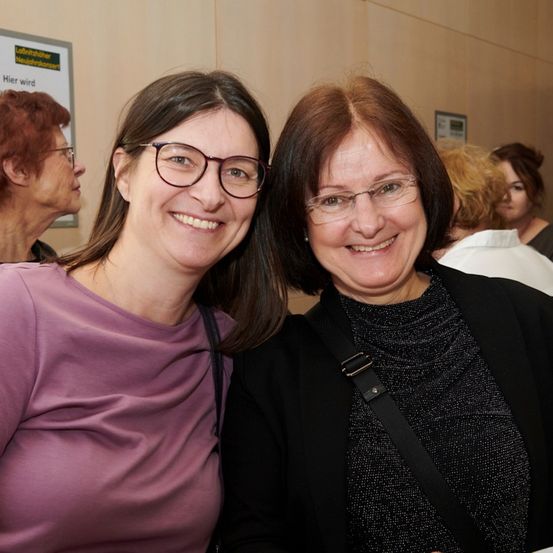 Zwei Frauen lächeln für ein Foto in einem Konferenzraum, mit anderen im Hintergrund. Eine trägt ein lila Shirt, die andere einen schwarzen Mantel. Ein Schild an der Wand steht 'Hier ist...'.