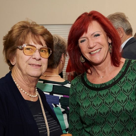Zwei Frauen lächeln, eine mit Brille und einer Halskette, die andere in einem grün gemusterten Shirt, stehen zusammen bei einer Veranstaltung mit anderen im Hintergrund.