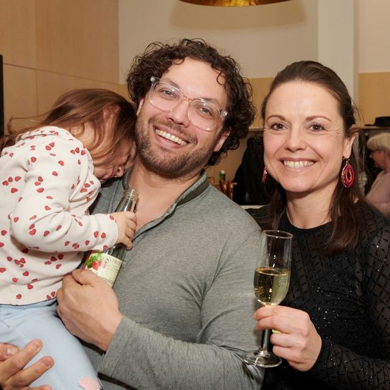 Ein lächelnder Mann hält ein Kind auf seiner Schulter, während eine Frau neben ihm ein Glas Champagner hält. Sie scheinen für ein Foto in einem Raum zu posieren.