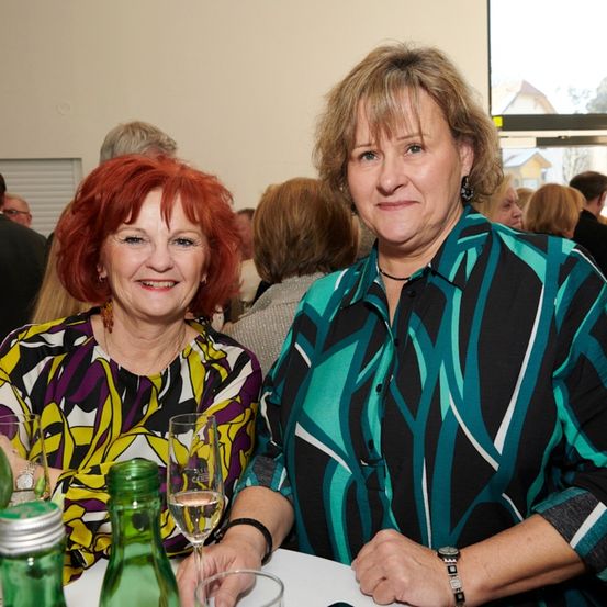 Zwei Frauen, eine mit roten Haaren und eine mit blonden, lächeln zur Kamera bei einer Veranstaltung in Innenräumen. Ein Weinglas und eine Flasche stehen auf dem Tisch vor ihnen.