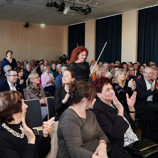 Eine Geigerin spielt vor einem Publikum in einem Saal. Das Publikum klatscht.